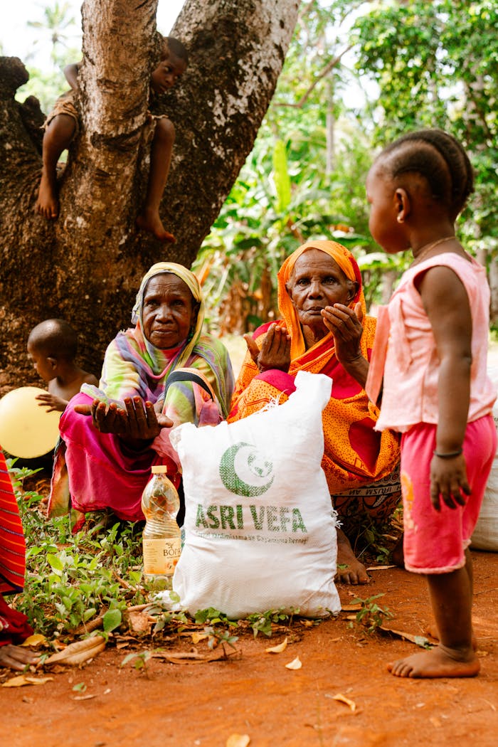 our-story Women and children gather under a tree with a bag labeled Asri Vefa, depicting a communal scene.